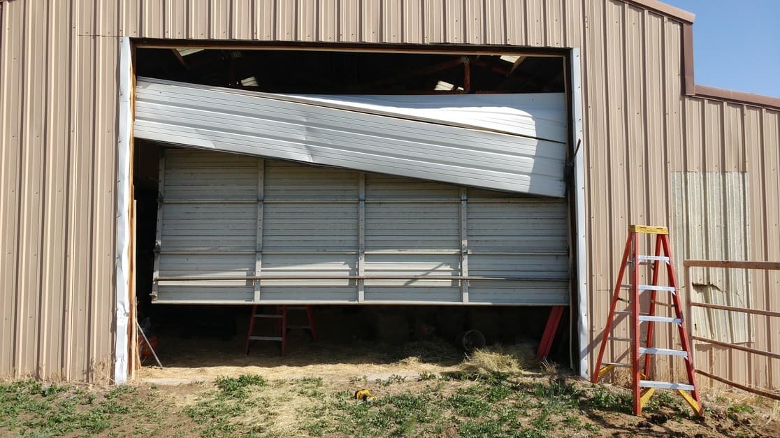 A metal barn's large garage door, its top section bent and twisted after being hit